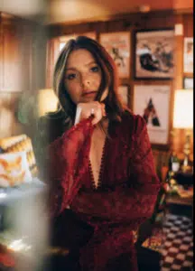 A woman in a red lace dress stands indoors, resting her chin on her hand, as if lost in thought, with framed pictures and warm lighting suggesting there’s a story behind the moment.